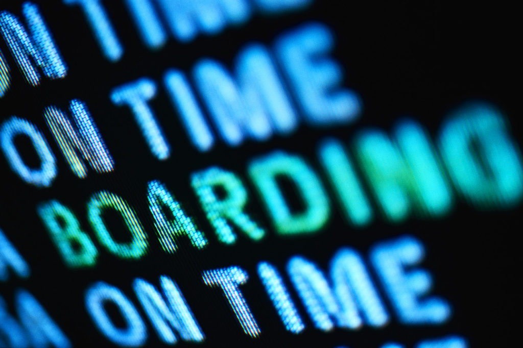 An airport terminal computer monitor shows the status of departing flights, some on time and one boarding.